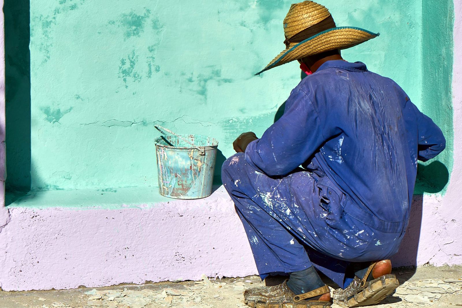 Pintor de chapéu de palha, agachado, pintando de verde-água a parede de uma casa, com a lata ao lado