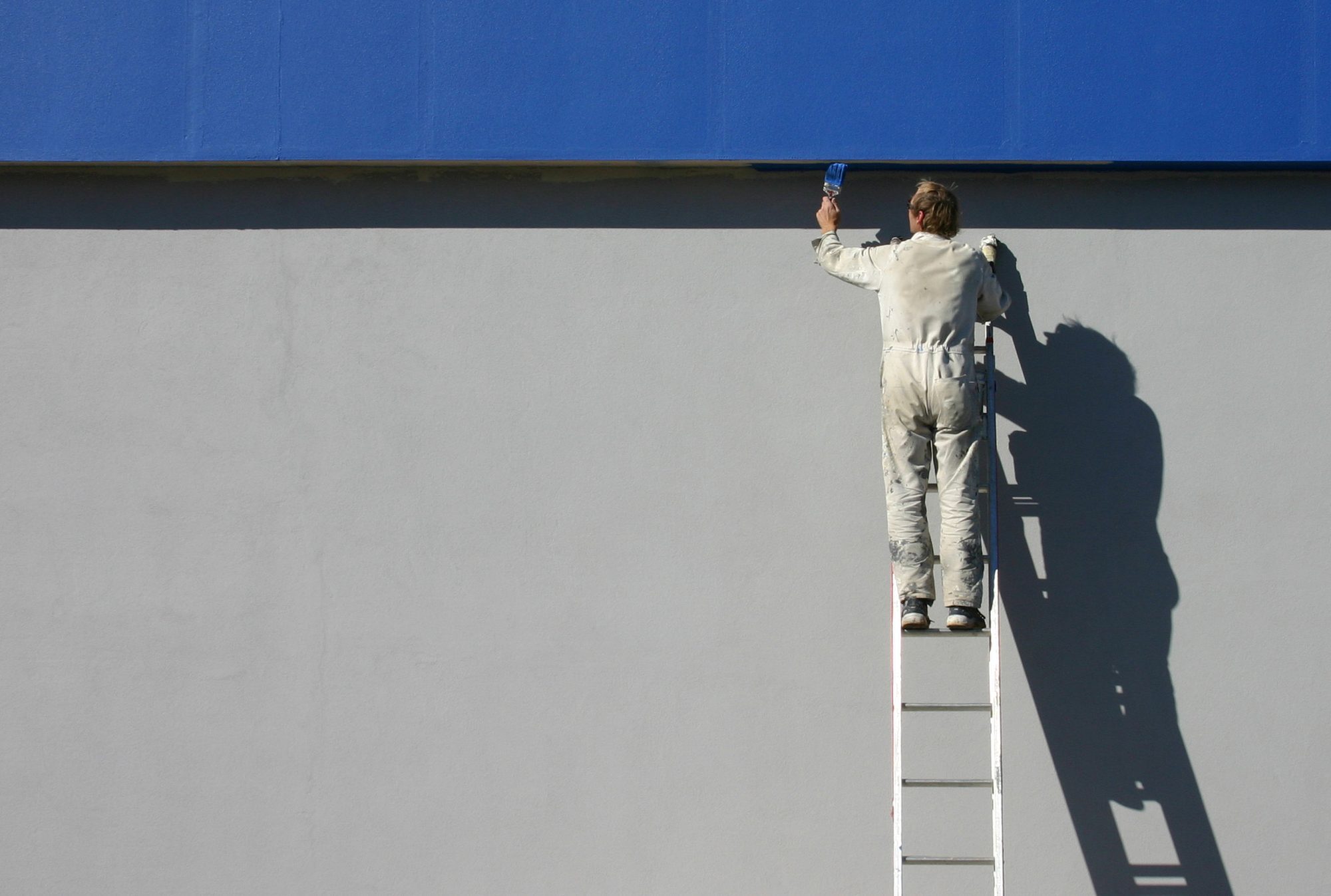 Pintor no alto de uma escada pintando uma parede cinza logo abaixo de uma faixa azul