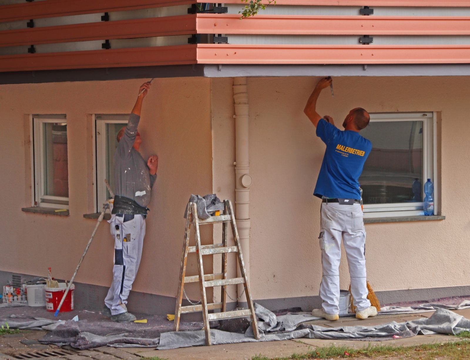 Dois pintores de branco pintando a fachada de uma casa, com a lona estendida no chão e uma escada aberta