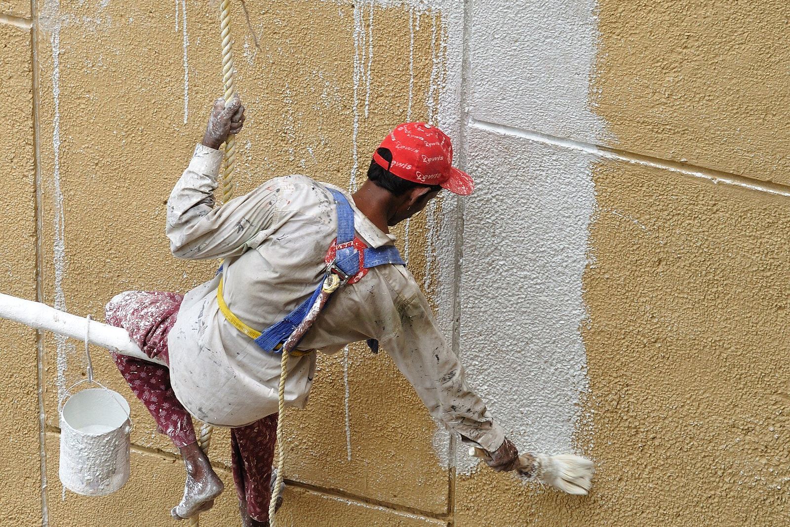 Pintor pendurado por cordas pintando de branco a fachada bege de um prédio
